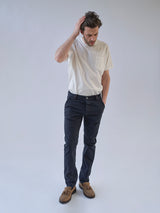 A man wearing a white t-shirt, Blue de Gênes BGpaulo Pavia 1 V2 Chino in Midnight Blue, and brown shoes stands against a plain light background, looking down with one hand on his head and the other in his pocket.