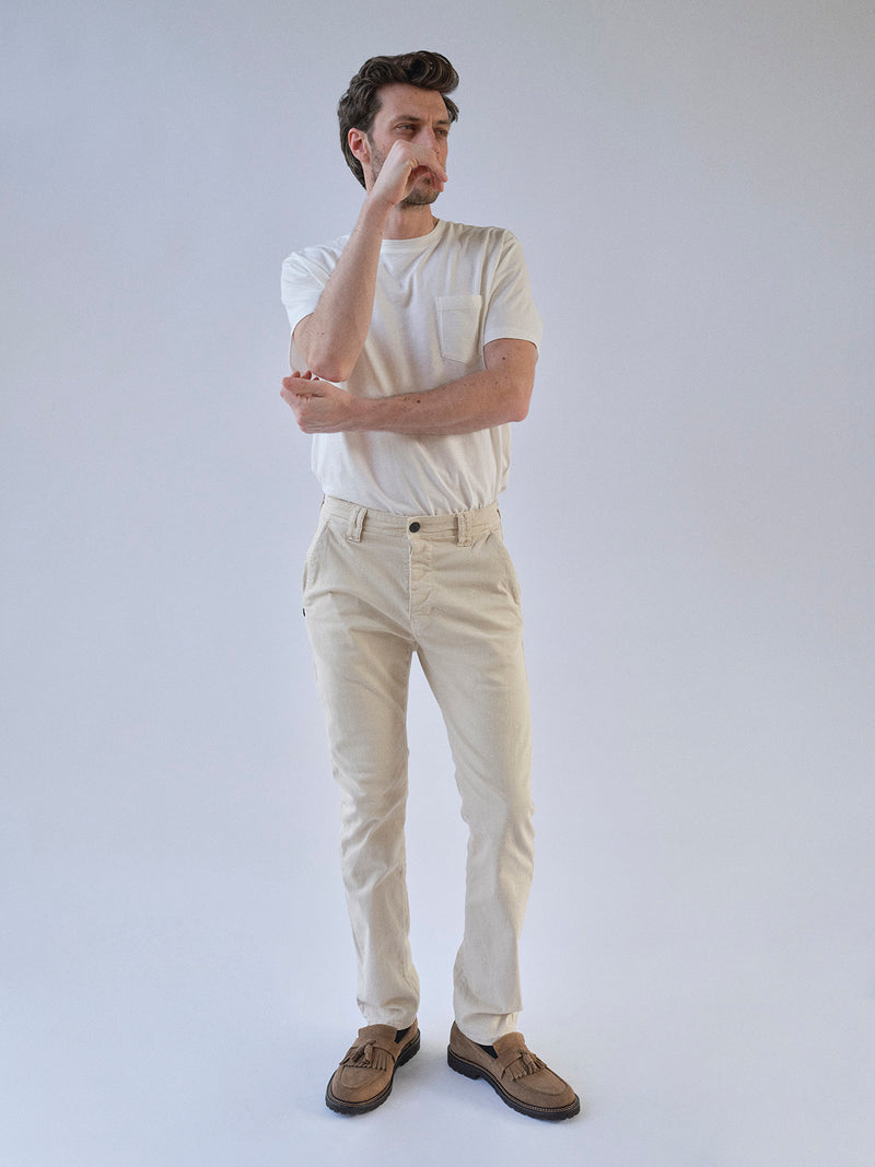 A man stands against a plain background, wearing a white stretch cotton t-shirt and Blue de Gênes BGpaulo Pavia 1 V2 Chino pants in ecru, with brown shoes. He crosses one arm and touches his face, gazing thoughtfully into the distance.