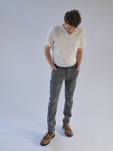A man in a white t-shirt, Blue de Gênes Repi Giulio Light V2 Jeans in grey denim, and brown shoes stands against a plain light background, looking down with his hands in his pockets.