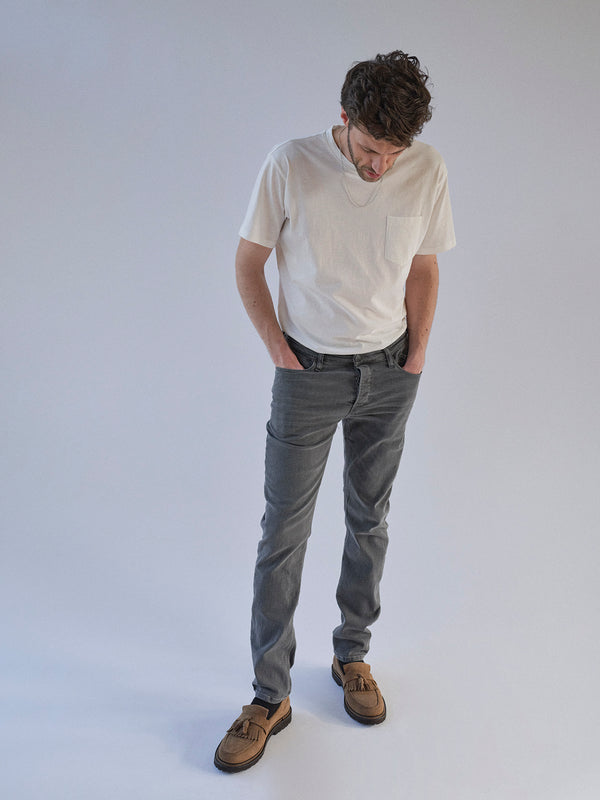 A man in a white t-shirt, Blue de Gênes Repi Giulio Light V2 Jeans in grey denim, and brown shoes stands against a plain light background, looking down with his hands in his pockets.