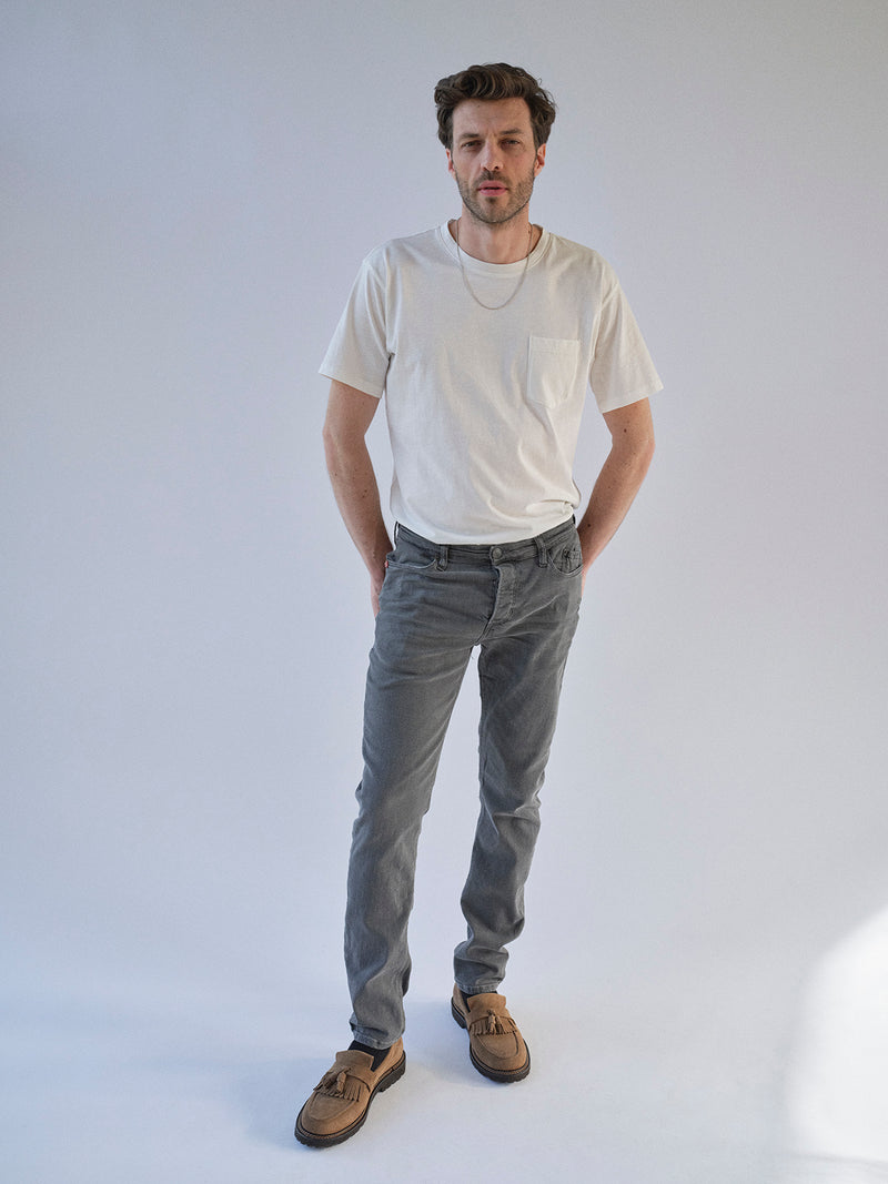 A man with short brown hair and a beard stands against a plain background, wearing a white T-shirt, Blue de Gênes Repi Giulio Light V2 Jeans in grey denim, brown loafers, and a silver chain necklace. His hands are in his pockets as he faces the camera.
