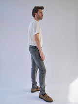 A man stands in profile against a plain light background, wearing a white t-shirt, Blue de Gênes Repi Giulio Light V2 Jeans in grey denim, and brown shoes. His hands are by his sides and he looks ahead with a neutral expression.