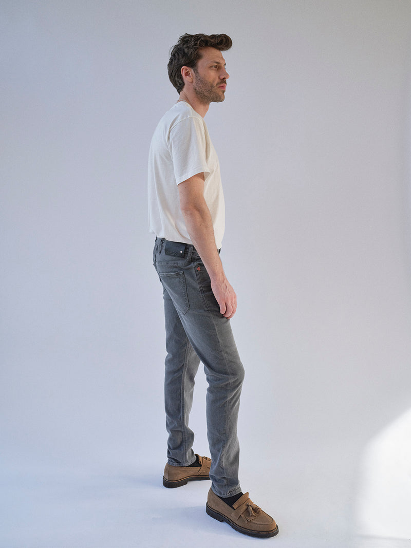 A man stands in profile against a plain light background, wearing a white t-shirt, Blue de Gênes Repi Giulio Light V2 Jeans in grey denim, and brown shoes. His hands are by his sides and he looks ahead with a neutral expression.