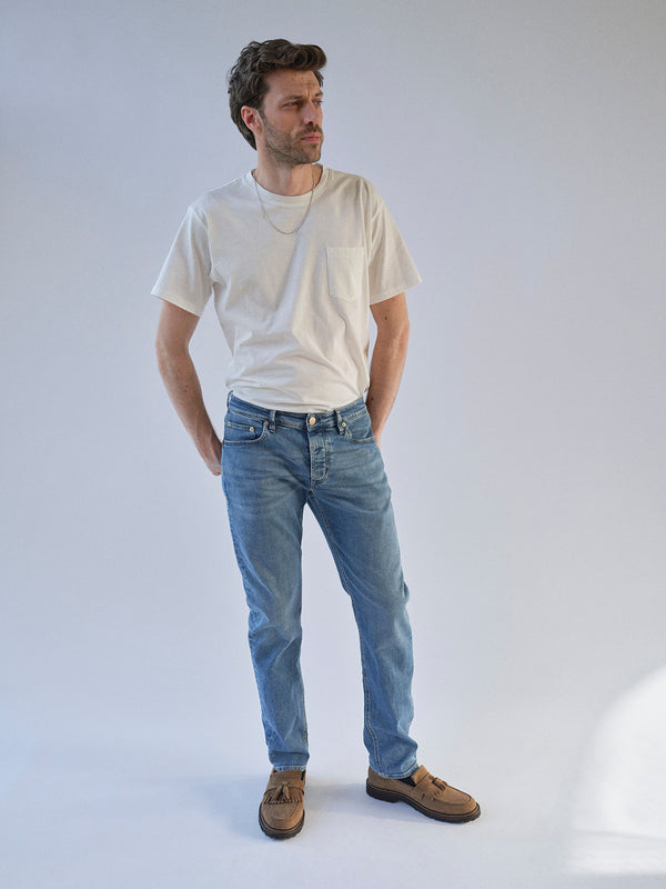 A man with short brown hair and a beard stands against a light background, wearing a white T-shirt, Blue de Gênes BGvinci Pala Used V2 Jeans in denim blue, a necklace, and brown shoes. His hands are in his pockets as he looks to the side.