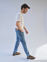 A man in a white t-shirt, BGvinci Pala Used V2 Jeans – Denim Blue by Blue de Gênes, and brown loafers walks sideways in a brightly lit studio with a plain light background.