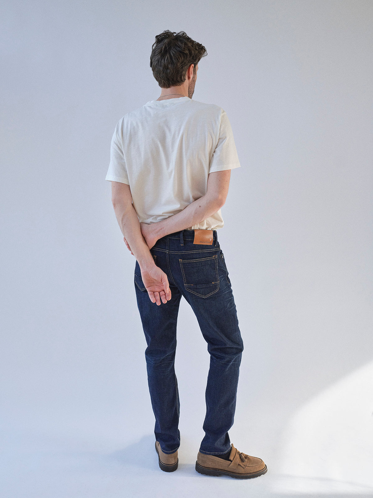 A man stands with his back to the camera, wearing a white T-shirt, Blue de Gênes BGvinci Pala Dark V2 Jeans in dark blue denim, and brown shoes. One arm is bent behind his back as he holds his opposite wrist.