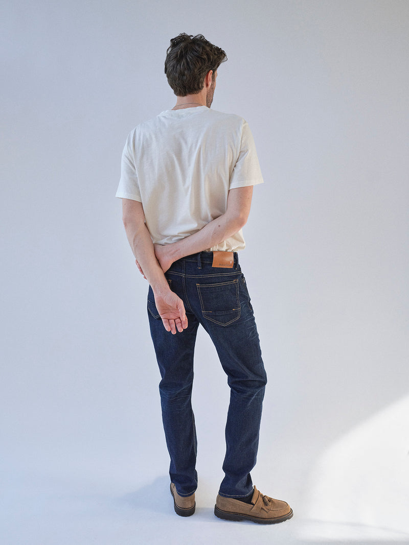 A man stands with his back to the camera, wearing a white T-shirt, Blue de Gênes BGvinci Pala Dark V2 Jeans in dark blue denim, and brown shoes. One arm is bent behind his back as he holds his opposite wrist.
