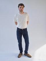 A man stands against a plain light background, wearing a white t-shirt, Blue de Gênes BGvinci Pala Dark V2 Jeans in dark blue denim, brown shoes, and a necklace. His hands are behind his back as he looks slightly to the side.