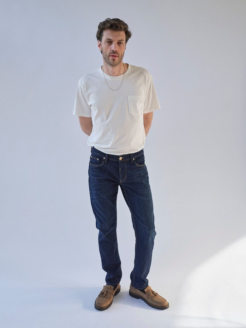 A man stands against a plain light background, wearing a white t-shirt, Blue de Gênes BGvinci Pala Dark V2 Jeans in dark blue denim, brown shoes, and a necklace. His hands are behind his back as he looks slightly to the side.