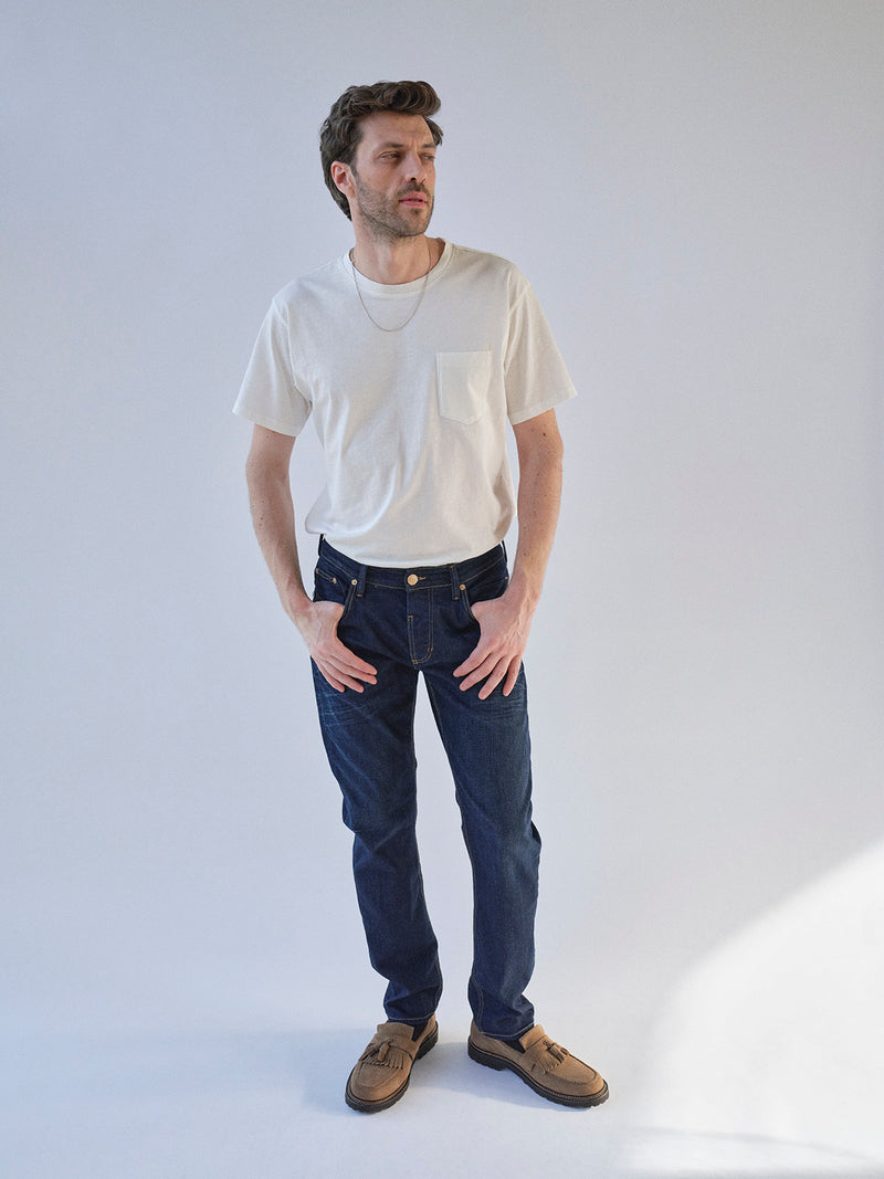 A man wears a white T-shirt, Blue de Gênes BGvinci Pala Dark V2 Jeans in dark blue denim, and brown shoes. He stands against a light background, looking slightly to the side with his hands in his pockets.
