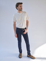 A man stands in a well-lit room, wearing a white t-shirt, brown shoes, and Blue de Gênes BGrepi Dai V Dark V2 Jeans in dark blue denim. He looks to the side with one hand by his side. The background is plain and light-colored.