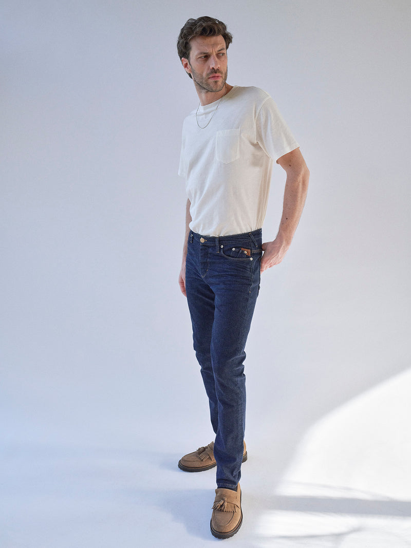 A man wearing a white t-shirt, Blue de Gênes BGrepi Dai V Dark V2 Jeans in dark blue denim, and brown shoes stands against a light background, looking to his left with one hand in his pocket.