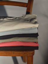A neatly folded stack of BGoscar T-Shirts by Blue de Gênes, featuring the timeless crewneck design in assorted colors like white, blue, olive green, red, and black—including the signature Dark Navy—rests on a wooden chair. Each shirt is crafted from soft 100% cotton for everyday comfort.