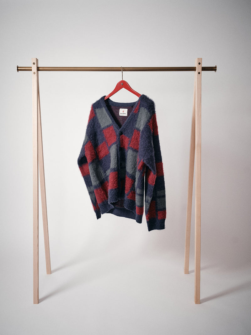 The Blue de Gênes Block Knit Cardigan - Navy Pattern, featuring a bold check design, hangs on a red hanger from a minimalist wooden rack against a plain white background.