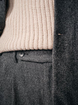 Close-up of a person wearing a textured beige knit sweater tucked into Blue de Gênes Kyoto Pant in black pattern, with a matching gray herringbone coat partially visible.