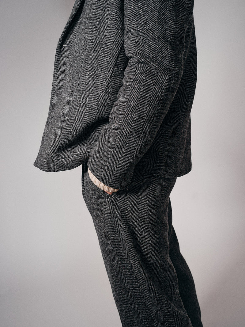 A person, shown from shoulders down against a plain background, wears refined tailoring—a grey herringbone suit with Blue de Gênes Kyoto Pant in Black Pattern—and has their hand in their pocket.