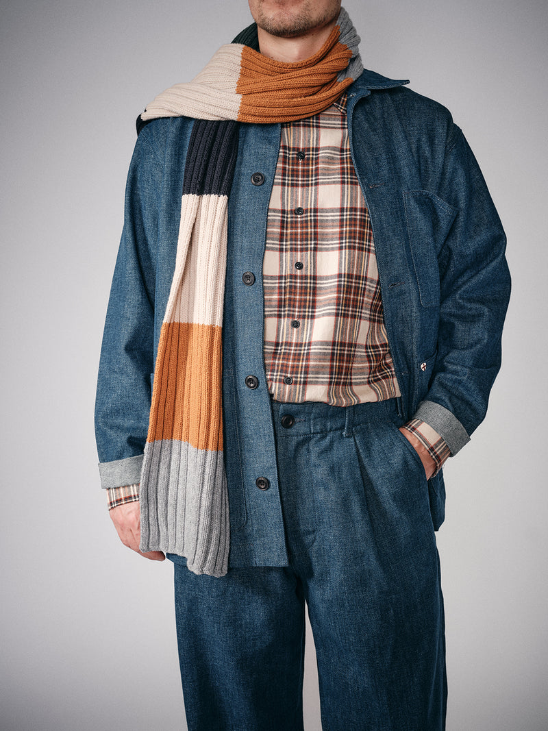 A man in a blue denim jacket and pants over a plaid shirt wears the Blue de Gênes Comforto Knit Scarf - Multi Stripe, featuring blocks of orange, gray, cream, and navy. His face is partially out of frame.