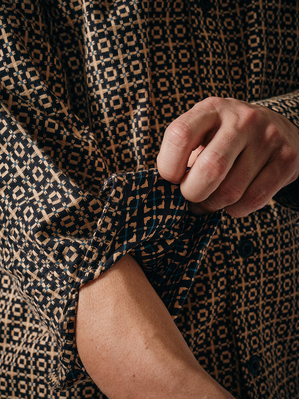 A person rolls up the sleeve of the Blue de Gênes Manga Overshirt – Black Pattern, showcasing its geometric black and beige checkered design in a close-up that highlights both hand and this versatile staple.