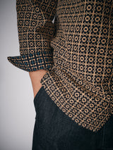 A person stands with one hand in their pocket, wearing the Blue de Gênes Manga Overshirt - Black Pattern, a patterned brown and black long-sleeve cotton shirt with rolled cuffs and a bold geometric square design, paired with dark denim jeans.