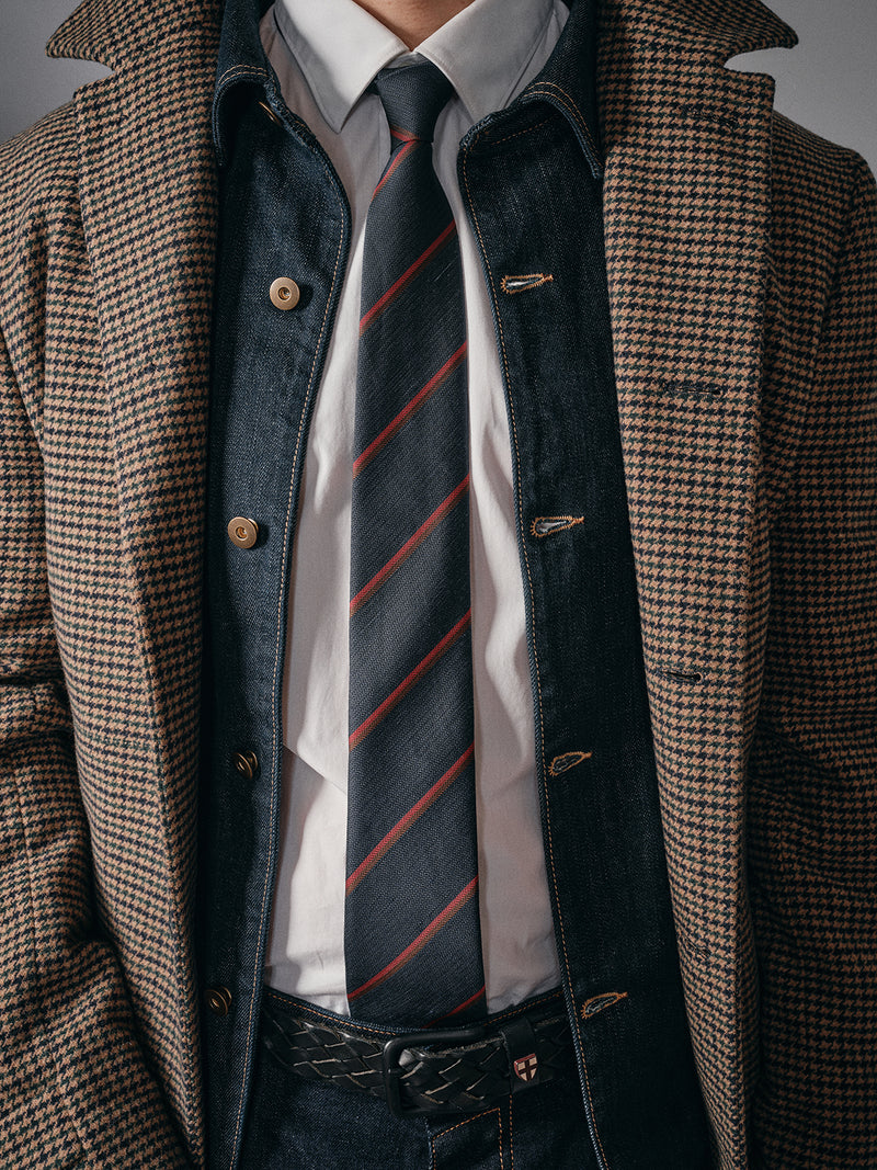 A person wears a houndstooth coat over a denim jacket, white dress shirt, dark jeans with a black belt, and finishes the look with Blue de Gênes’ Nekutaki Tie #B - Navy Stripe, blending formal and casual styles in timeless mens fashion.