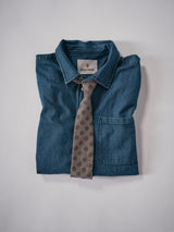 A folded blue denim shirt with a front pocket from Blue de Gênes, topped with the Nekutaki Tie #E in a sand pattern, both arranged neatly against a plain white background.