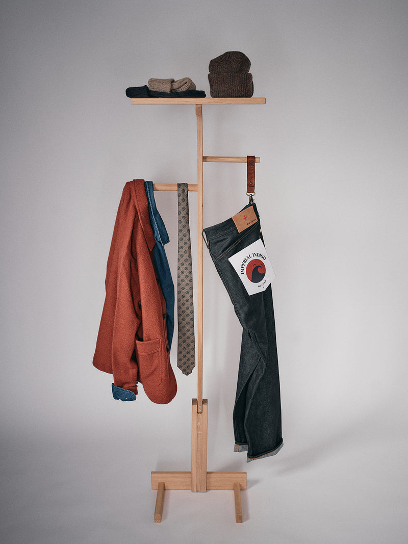 A wooden coat rack displays a red jacket, Nekutaki Tie #E – Sand Pattern by Blue de Gênes, belt, folded jeans with Made in France label, brown beanie, and gloves—celebrating tailoring traditions against a plain background.