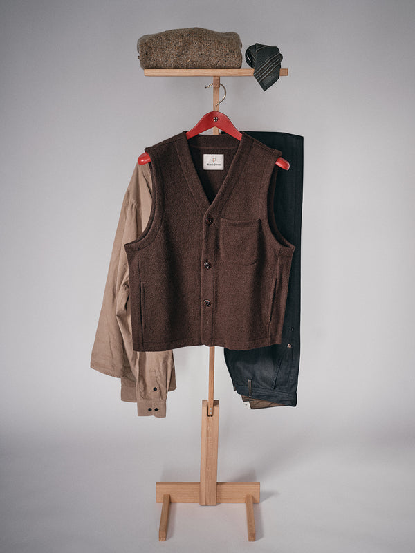 A wooden valet stand displays a Blue de Gênes Koi Wool Gilet - Chocolate vest, tan coat, dark trousers, a folded brown sweater on top, and a dark tie draped over the upper hook, set against a plain light background.