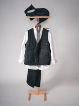 A Maurice Solid Gilet - Dark Navy by Blue de Gênes is displayed child-sized on a wooden stand with a white shirt, gray plaid tie, dark trousers, and a matching cap and pouch above, set against a plain neutral background.