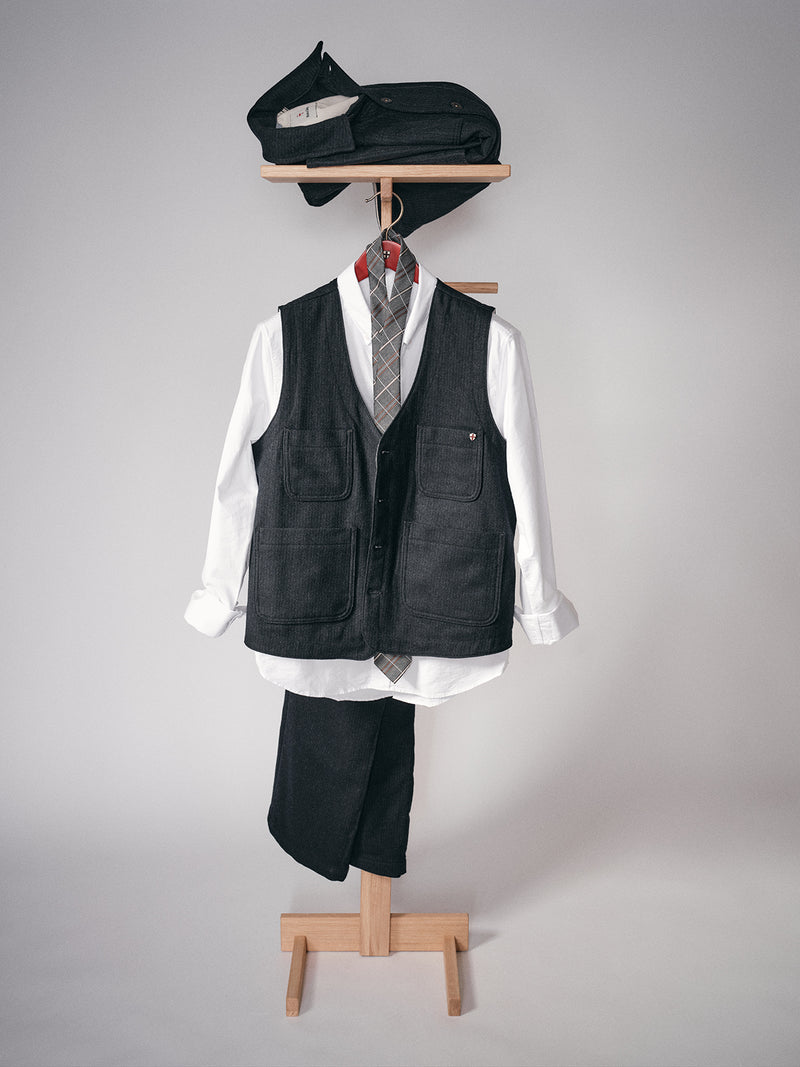 A Maurice Solid Gilet - Dark Navy by Blue de Gênes is displayed child-sized on a wooden stand with a white shirt, gray plaid tie, dark trousers, and a matching cap and pouch above, set against a plain neutral background.