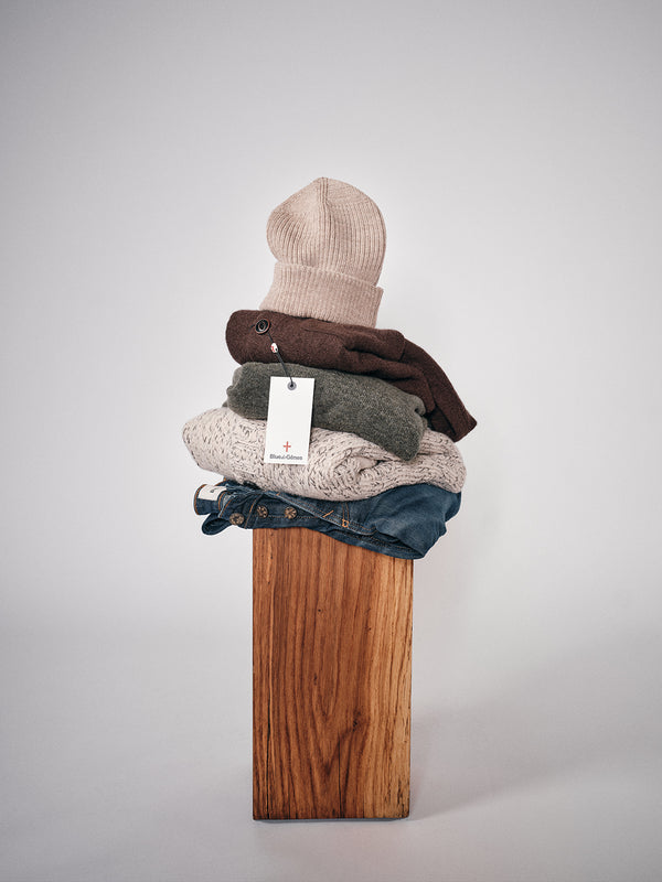 A stack of folded clothing, featuring the Boshi Beanie - Oat by Blue de Gênes, brown and gray sweaters (one with a white tag), and blue jeans, rests on a wooden pedestal against a plain light background.
