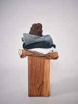 A neatly stacked set of clothes features the Boshi Beanie - Brown by Blue de Gênes, along with a blue sweater, dark speckled sweater, white shirt, and tan pants on a wooden pedestal set against a plain gray background.