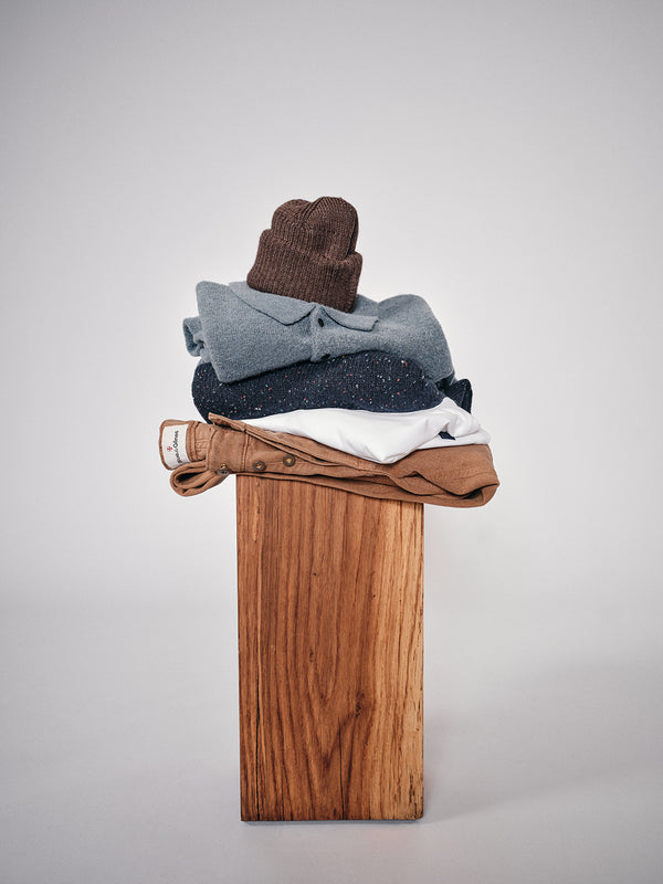 A neatly stacked set of clothes features the Boshi Beanie - Brown by Blue de Gênes, along with a blue sweater, dark speckled sweater, white shirt, and tan pants on a wooden pedestal set against a plain gray background.