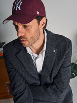 A man in a maroon New York Yankees cap, white shirt, and the Blue de Gênes Fukui Blazer – Black Pattern gazes to the side with a serious expression. The blazer’s relaxed tailoring is highlighted, with a plant and books in the background.