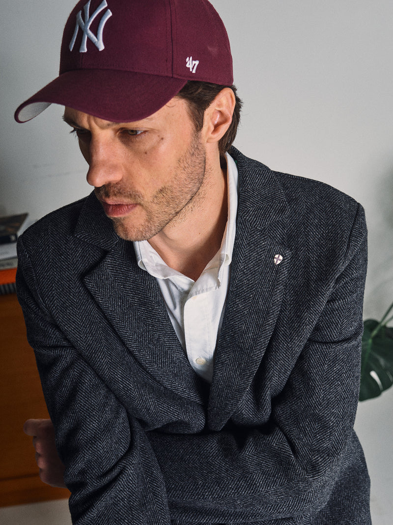 A man in a maroon New York Yankees cap, white shirt, and the Blue de Gênes Fukui Blazer – Black Pattern gazes to the side with a serious expression. The blazer’s relaxed tailoring is highlighted, with a plant and books in the background.