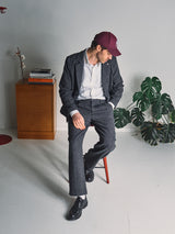 A man in the Blue de Gênes Fukui Blazer – Black Pattern, a white shirt, and a maroon baseball cap sits on a stool, looking down. Behind him are a wooden cabinet with books and a plant. The setting is minimal and modern.