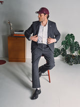 A man in a gray suit, white shirt, maroon cap, and Blue de Gênes Kyoto Pant - Black Pattern sits on a stool adjusting his jacket. Behind him are a wooden cabinet with books and a branch, plus a large green monstera plant.