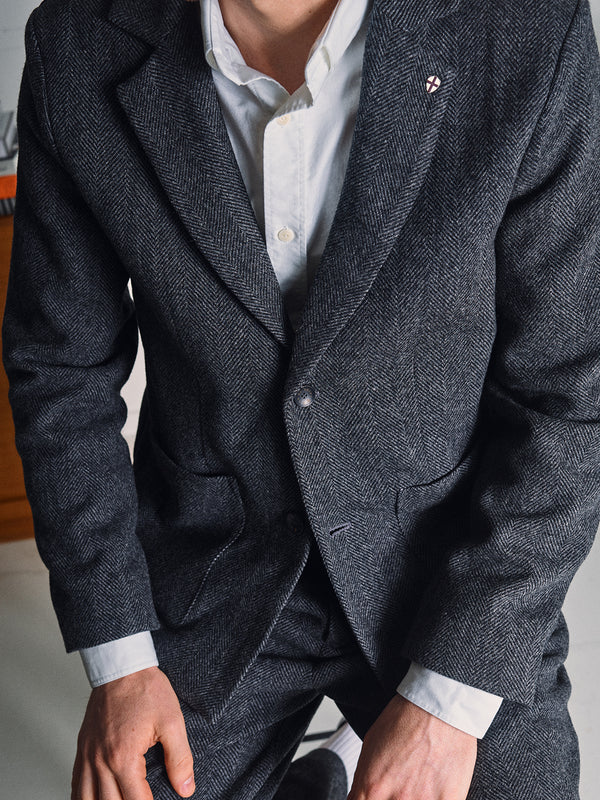 A person in a Blue de Gênes Fukui Blazer - Black Pattern with relaxed tailoring and a small lapel pin, over a white dress shirt, is kneeling with their face partially out of frame.