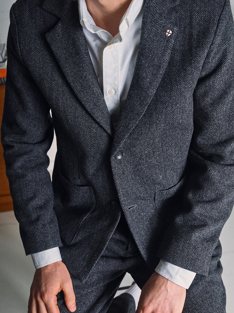 A person in a Blue de Gênes Fukui Blazer - Black Pattern with relaxed tailoring and a small lapel pin, over a white dress shirt, is kneeling with their face partially out of frame.