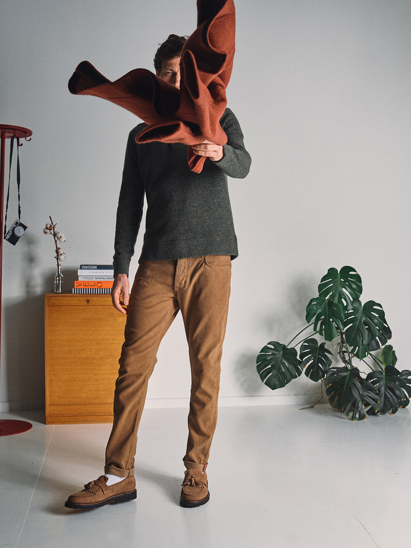 A man in brown pants and a Blue de Gênes Malakka Polo Knit - Army stands indoors, partially covering his face with a folded rust-colored coat. In the background are a wooden cabinet, books, plant, and coat rack.