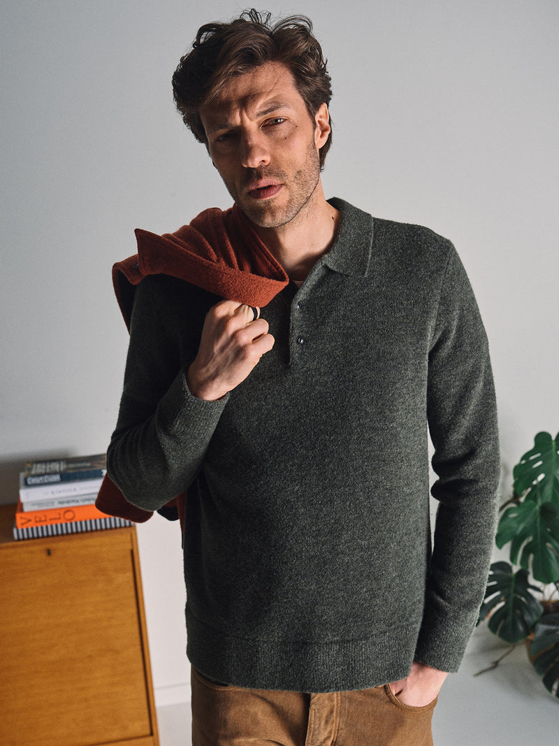 A man with wavy brown hair wears the Blue de Gênes Malakka Polo Knit - Army sweater with corozo buttons and tan pants, holding a rust jacket indoors near a wooden cabinet filled with books and a leafy green plant in the background.