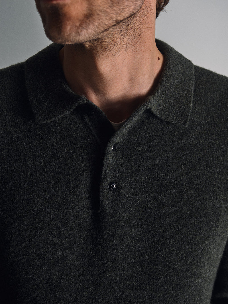 A close-up shows a man in the Blue de Gênes Malakka Polo Knit - Army, with corozo buttons, collar, and long sleeves. His neck, jawline, and stubble are visible against a plain background.