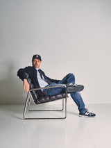 A man wearing the Blue de Gênes Gifu Knit Cardigan in navy, a black baseball cap, and jeans sits casually with legs crossed in a modern black and chrome chair against a white wall, his left arm resting on the chair.