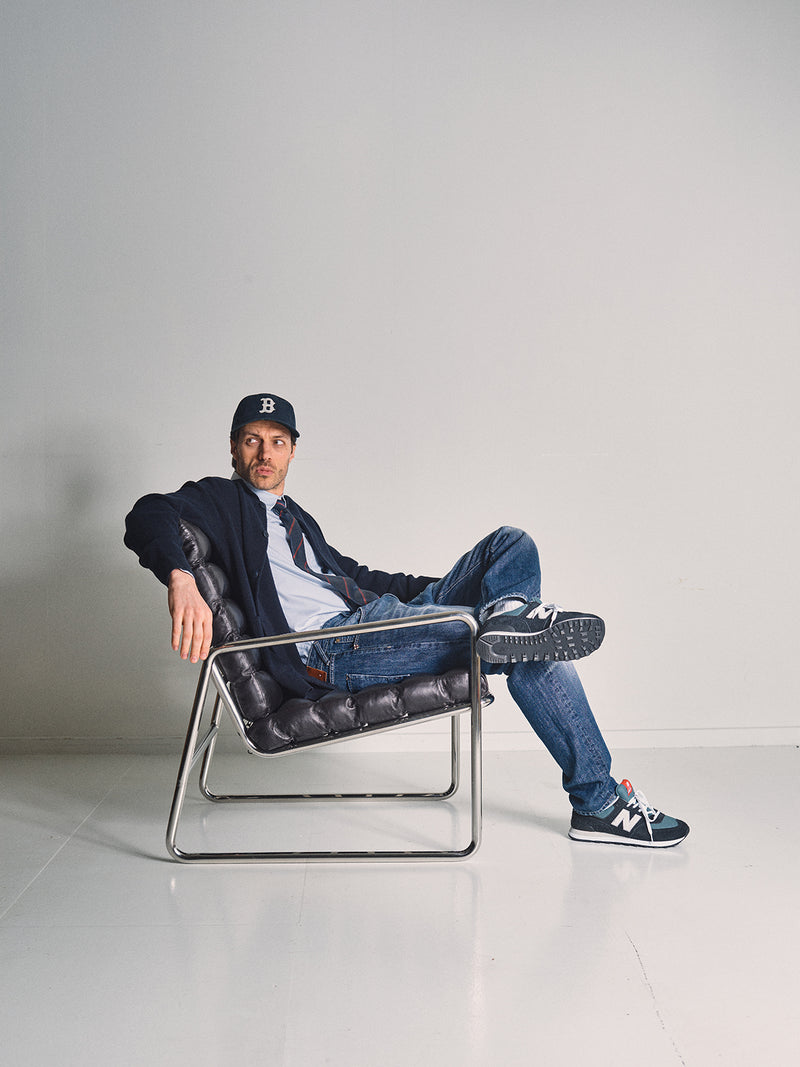 A man wearing the Blue de Gênes Gifu Knit Cardigan in navy, a black baseball cap, and jeans sits casually with legs crossed in a modern black and chrome chair against a white wall, his left arm resting on the chair.