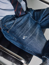 A person sits in a black leather chair wearing Blue de Gênes Giacomo Osaka Jeans in dark denim, made in Italy, with a relaxed loose fit and a small ripped patch on the left thigh. The focus is on the jeans.