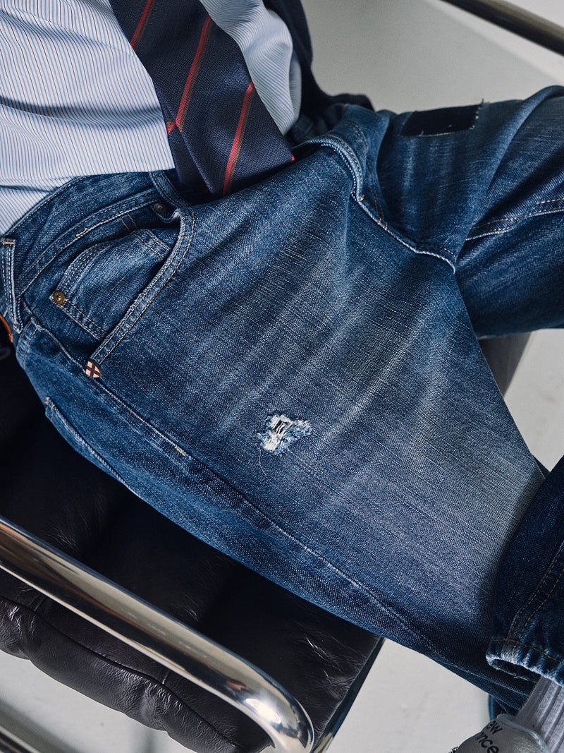 A person sits in a black leather chair wearing Blue de Gênes Giacomo Osaka Jeans in dark denim, made in Italy, with a relaxed loose fit and a small ripped patch on the left thigh. The focus is on the jeans.