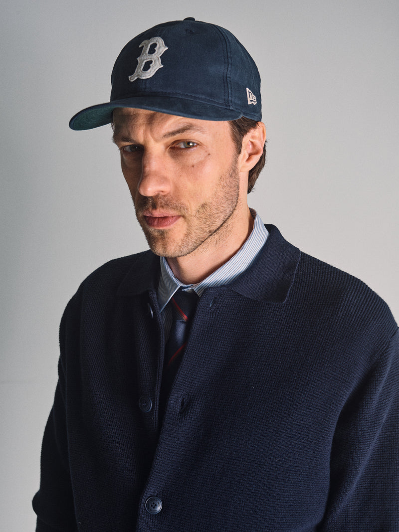 A man stands against a plain light background wearing the Blue de Gênes Gifu Knit Cardigan in navy, along with a baseball cap featuring a B logo, a blue striped shirt, and a tie.
