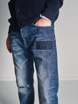 A person wearing Blue de Gênes Giacomo Osaka Jeans - Dark Denim, featuring a relaxed loose fit with a thigh patch and light knee distressing, paired with a dark jacket and light blue shirt. Only the lower torso and upper legs are visible.