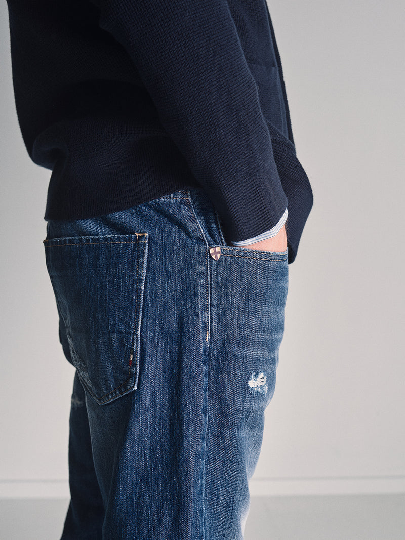 Wearing Blue de Gênes Giacomo Osaka Jeans in Dark Denim, a person faces sideways against a plain light background, one hand in their pocket and dressed in a navy blue sweater.