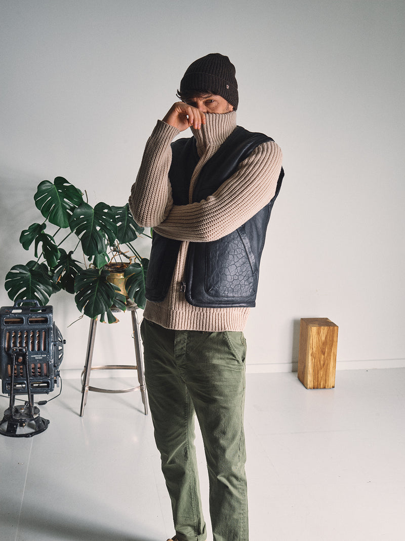 Indoors, a person in green pants, a black beanie, and the Blue de Gênes Saga Shearling Gilet - Scarab poses with one arm raised, wearing a beige turtleneck. A potted plant, small heater, and wooden block are visible behind them.
