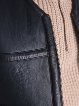Close-up of the Saga Shearling Gilet - Scarab by Blue de Gênes, featuring black leather and a visible zipper, worn over a beige textured knit sweater—crafted from premium German Merino sheepskin.